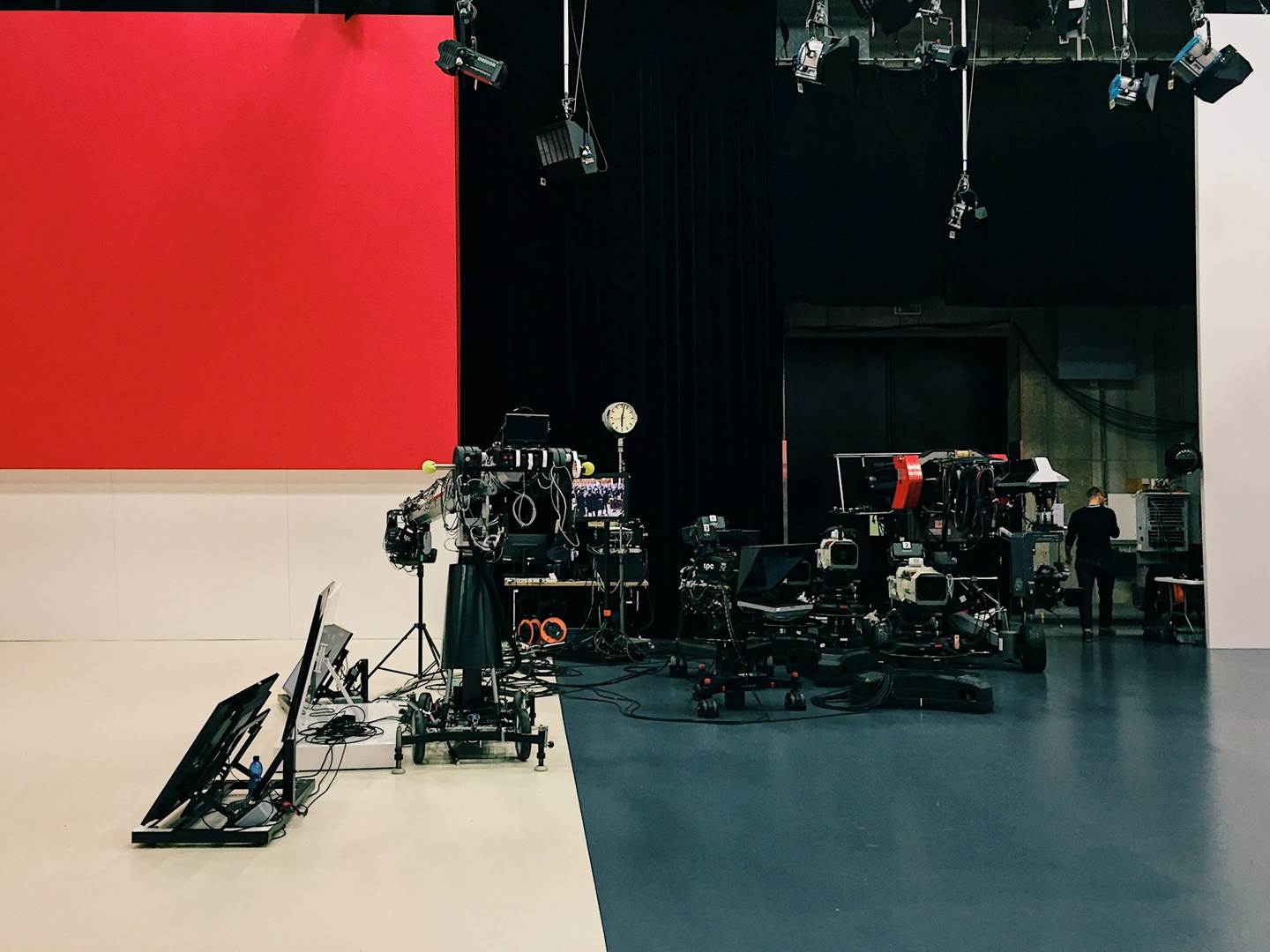 An empty television studio floor: a red cyclorama wall and white cove on the left, a lighting grid overhead, and a jib arm and a cluster of camera pedestals parked and waiting.
