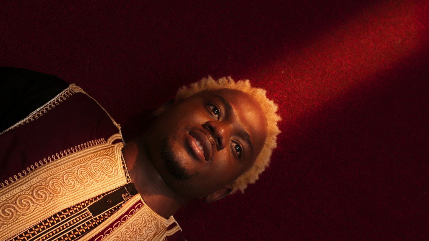 A young man with bleached curls lies back against deep crimson ground, an embroidered collar at his shoulder, looking straight up into the lens.