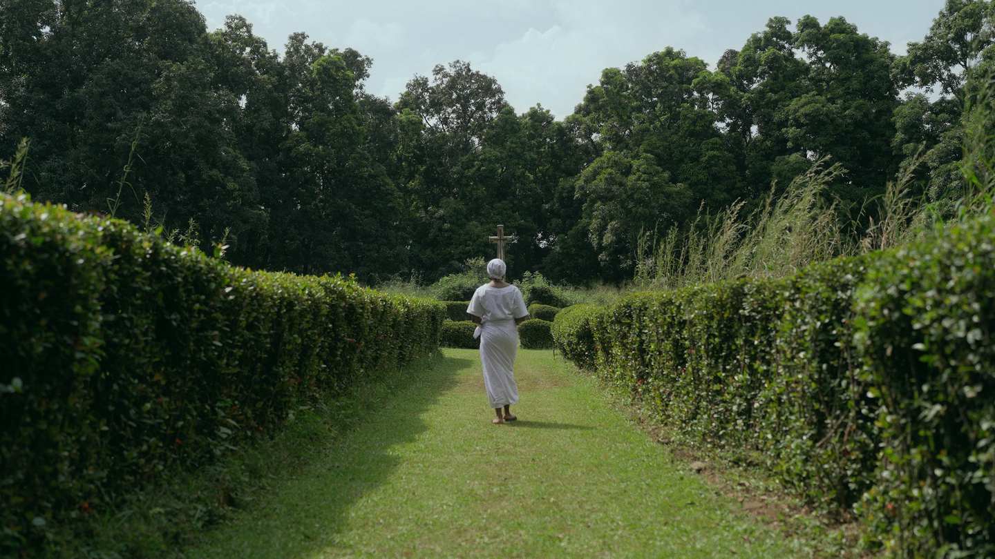 A figure in white robes walks away up a grass path between tall hedges, carrying a wooden cross.